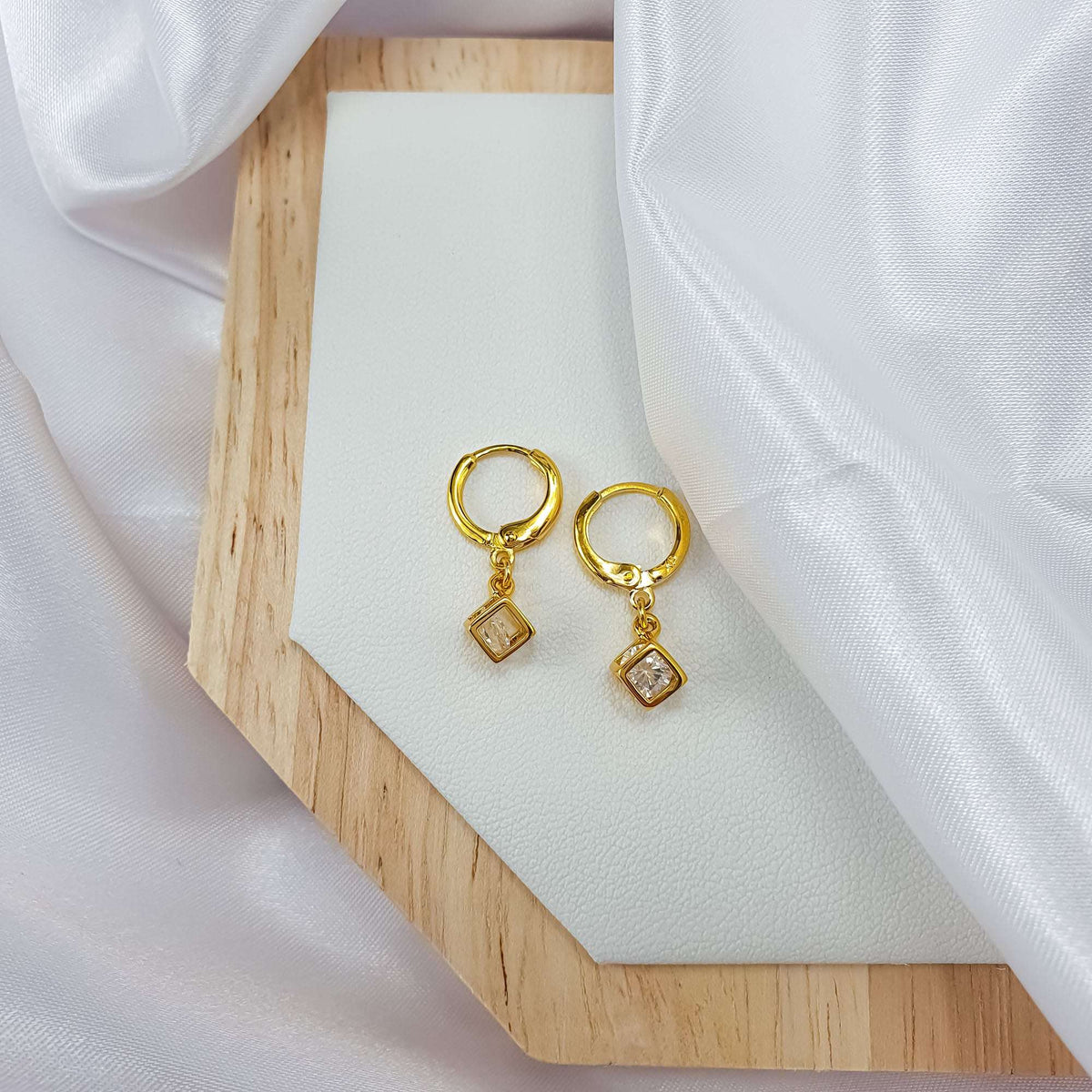 Paauksuotų žiedinių auskarų pora su kvadratiniais kristalo pakabukais ant medinio padėklo ir baltos šilko draperijos