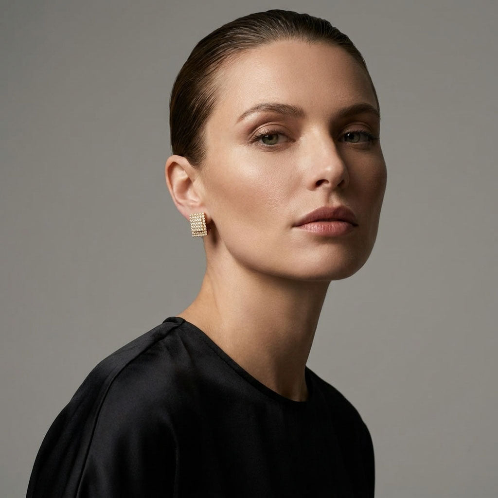 Modelė juoda palaidine ir žėrinčiais kvadratiniais cirkonių klipsais ausyje pilkame fone