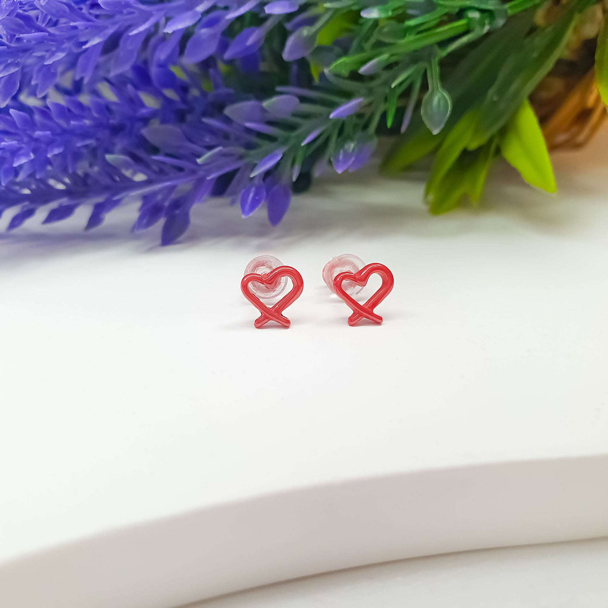 Raudonos emalio širdelių įsegami auskarai su skaidriais fiksatoriais baltame fone prie levandų